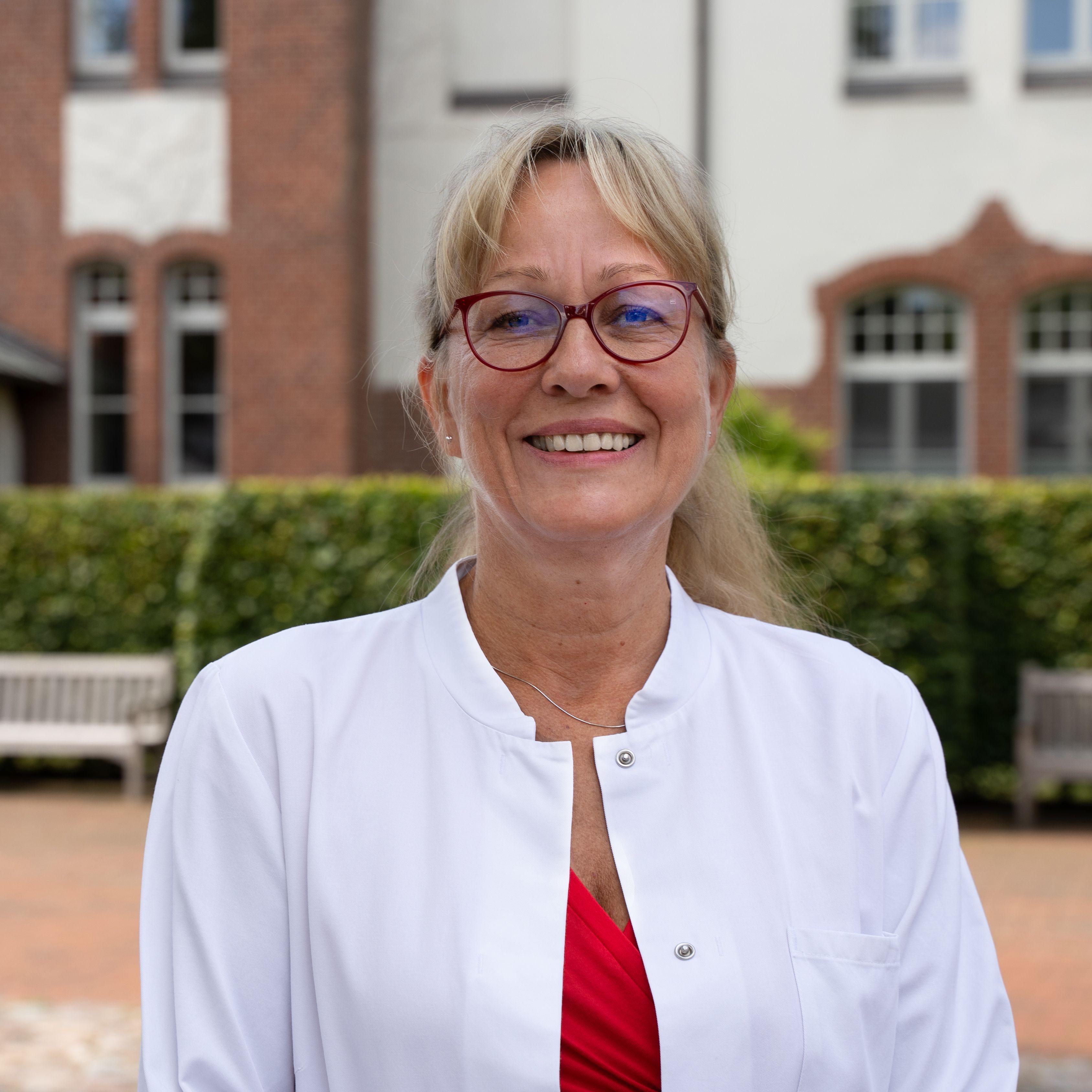 Frau Dr. Niecke steht vor der Rehaklinik Hohenelse. Sie trägt ein rotes Oberteil, eine schwarze Hose, darüber einen weißen Arztkittel. Sie hat blonde lange Haare und trägt eine rote Brille.