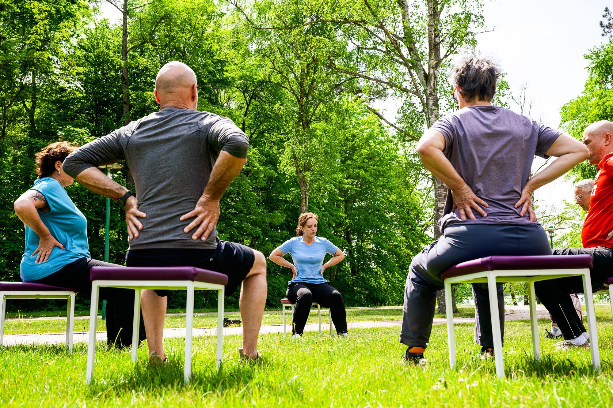 Patienten sitzen im Garten in einem Stuhlkreis und machen Übungen unter Anleitung einer Therapeutin.